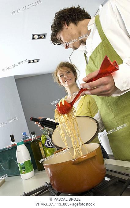 Side profile of a young man cooking noodles with a young woman smiling beside him