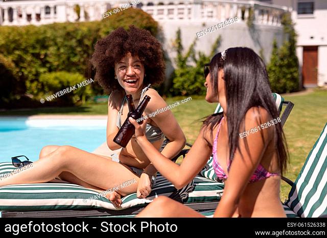 Two diverse female friends sunbathing by pool and drinking beer