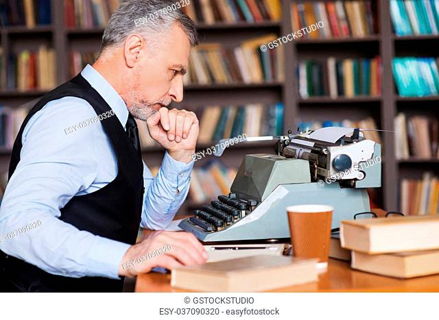Waiting for inspiration. Side view of confident grey hair senior man in formalwear sitting at the typewriter and holding hand on chin with bookshelf in the...