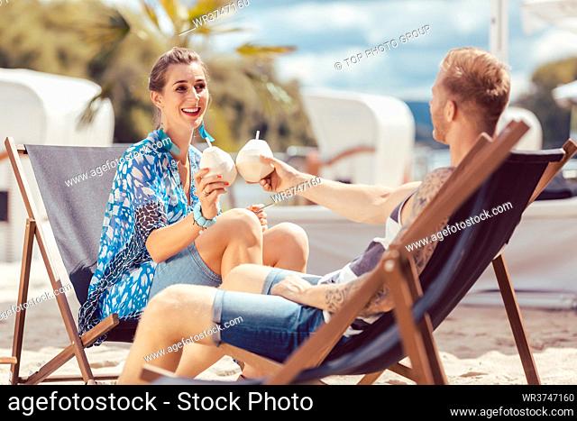 Young couple sitting on chair at beach toasting coconut water