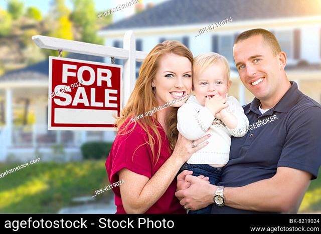 Happy young military family in front of for sale real estate sign and new house