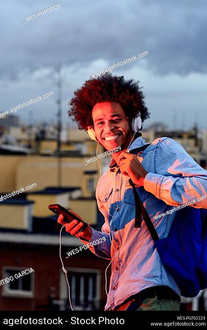 Smiling young man with backpack listening music over headphones against buildings in city