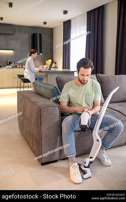 Vacuum cleaner setup. Young bearded man focused taking apart vacuum cleaner sitting on sofa and woman behind working on laptop