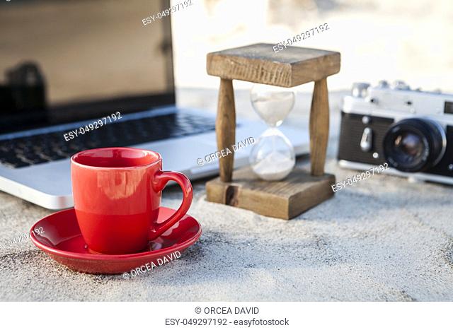 Red Coffe Mug and Vintage hourglass with silver laptop and old camera in the sand at the beach
