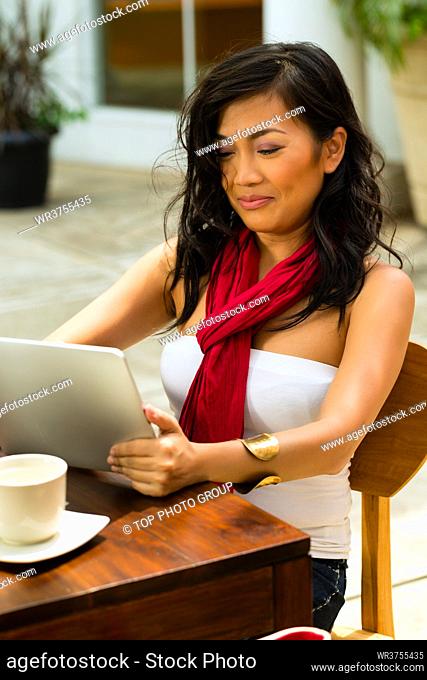 Asian woman is sitting in a bar or cafe outdoor and is surfing the internet with a tablet computer