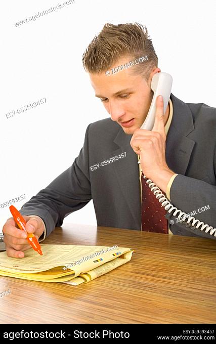 Young smiling businessman sitting at desk, talking on phone and marking something in newspaper. White background
