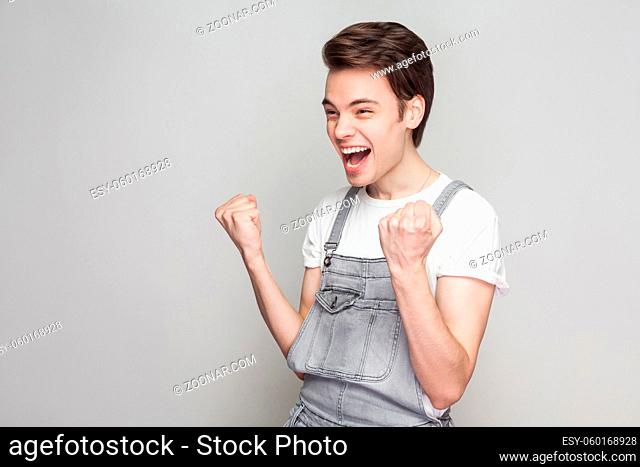 Portrait of happy amazed winner young brunette man in casual style with white t-shirt and denim overalls standing screaming and celebrating his victory
