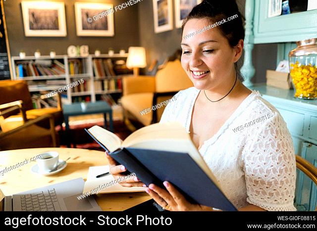 Smiling voluptuous woman reading book while sitting at table in restaurant