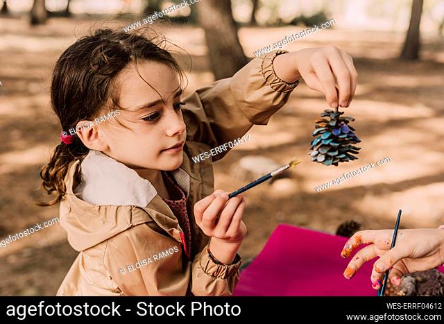 Cute girl holding pine cone while coloring by sister at park