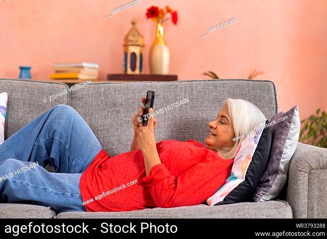 Old woman using mobile phone while lying down on sofa in living room