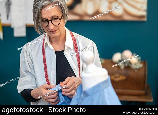 Tailor. Engaged serious middle aged woman in glasses standing near mannequin with pin touching blouse sleeve in sewing workshop