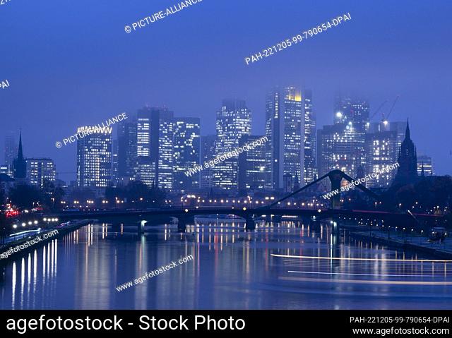 05 December 2022, Hessen, Frankfurt/Main: The tops of the bank towers of Frankfurt disappear in the fog, while a ship on the Main draws a light trail through...