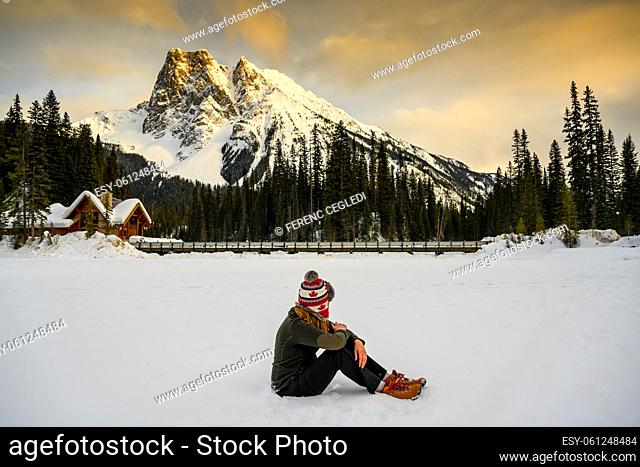 Casual woman sitting in the snow on the frozen Emerald Lake in the Yoho National Park, watching the sunset over Mount Burgess in the background close to Burgess...
