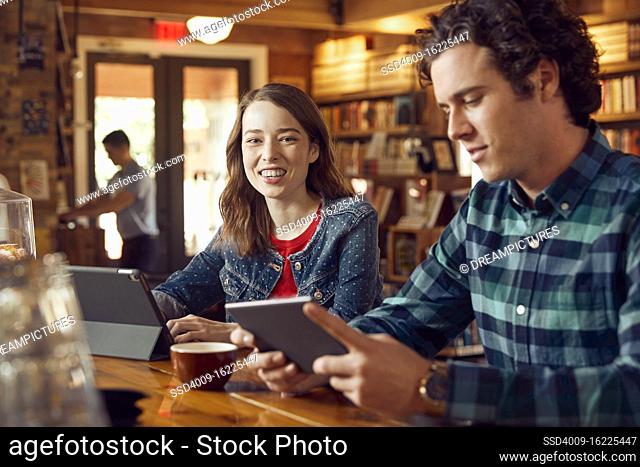 Young man and woman sitting at counter in cafe bookstore using digital tablets , woman looking at camera smiling