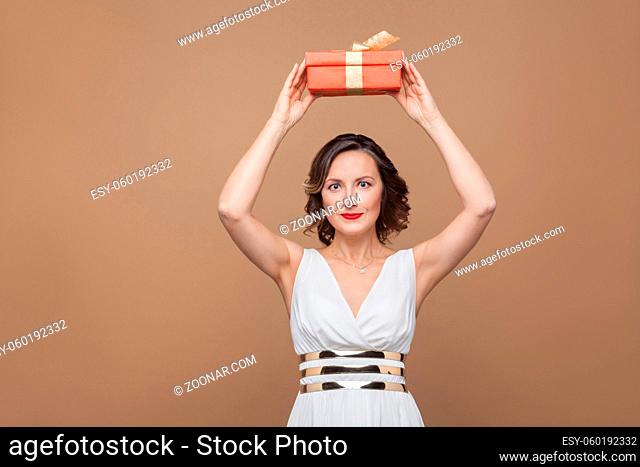 Beautiful brunette woman in white dress holding red gift box in top of head and looking at camera with smile. Emotion and feeling concept