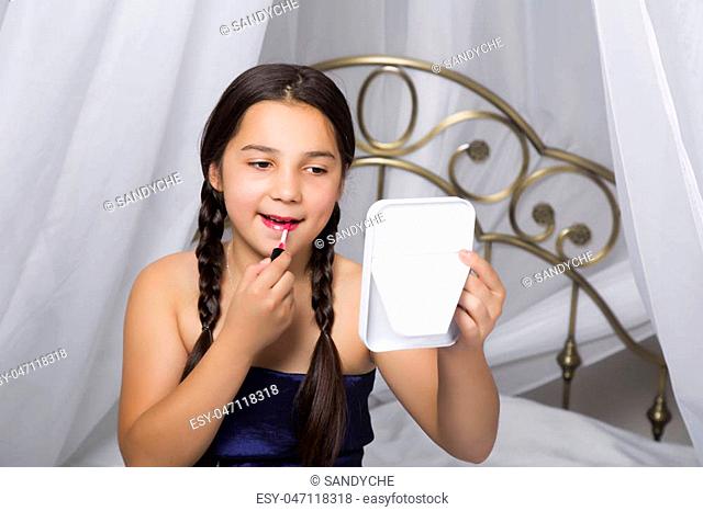 little girl sitting on a bed looks in the mirror and painting lips like her mom