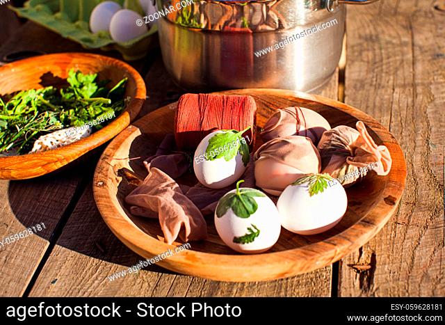 Close view wooden bowl with raw eggs in process of winding into synthetic material together with green plants, to create pattern after boiling in water with...