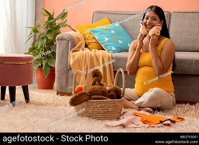 Smiling pregnant woman sitting on floor and holding a pair of baby shoes