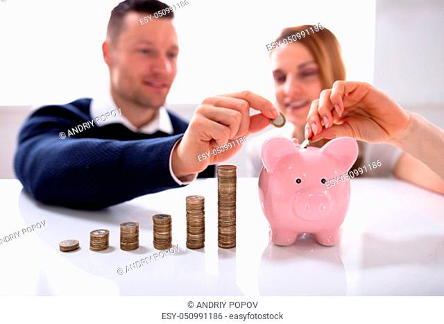 Couple Inserting Coin In Piggybank Near Increasing Profit Over Desk