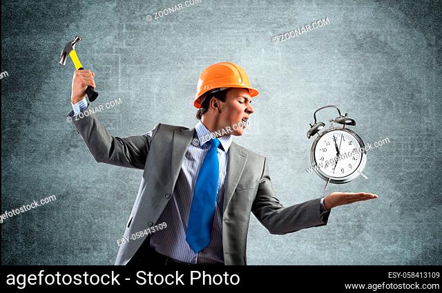 Businessman going to crash with hammer alarm clock. Young contractor in business suit and safety helmet standing on grey wall background