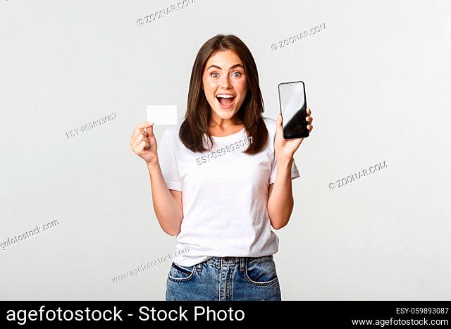 Excited and surprised cute girl showing credit card and mobile phone banking app on screen