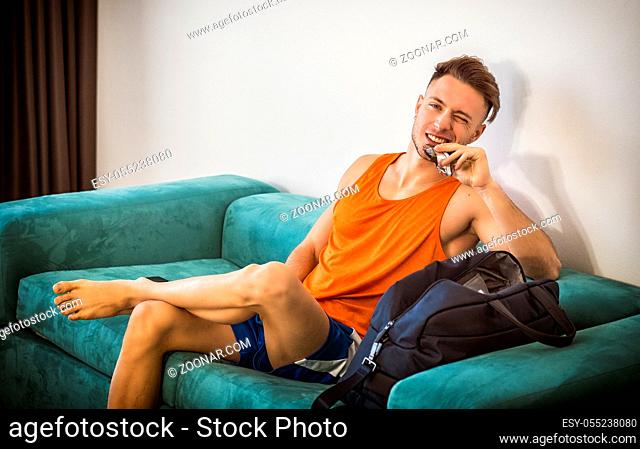 Young muscular man eating protein bar indoor at home sitting on a couch
