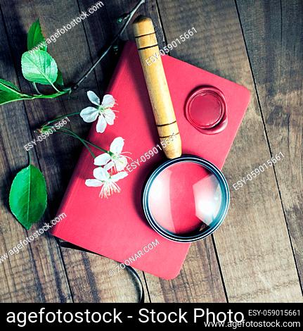 Red notebook, magnifier, wax seal, herry flowers and green leaves on vintage wood table background