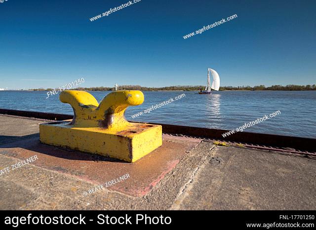 Sailing yacht under spinnaker on the ELbe. In the foreground a yellow steel bollard at the jetty Hamburg-Wittenbergen