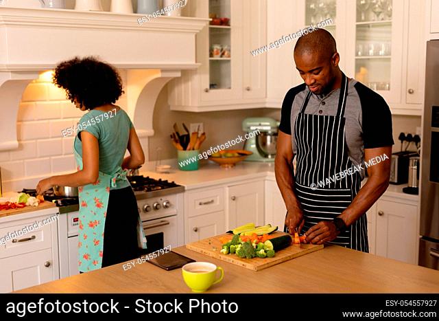 Front view of a mixed race couple at home standing in the kitchen wearing aprons preparing food, the man chopping vegetables and woman stirring a pot on the hob
