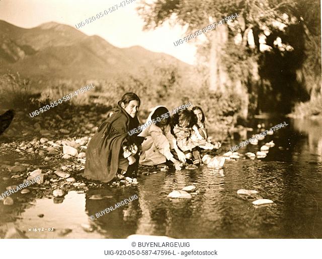 Four Taos children squat on rocks at edge of stream, mountains in background