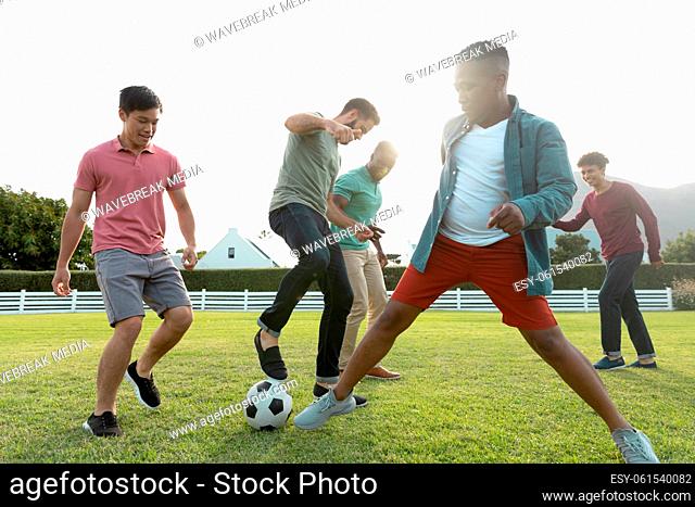 Multiracial male friends playing soccer on grassy land in yard against clear sky during weekend