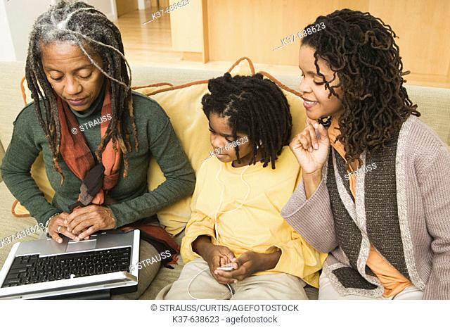 3 generations of African American family at home, with grandma using laptop being advised by older daughter & her grandson listening to his ipod