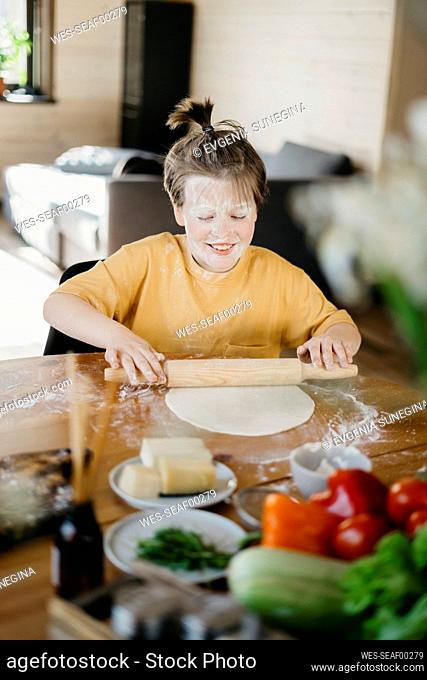 Smiling boy with flour on face rolling pin on dough at home