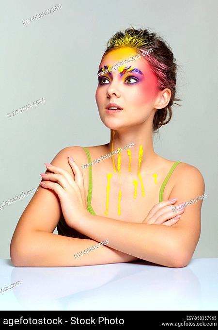 Female portrait with unusual face art make-up. Paint on brows, hair, around eyes and with paint drips on the neck. Woman sitting at a white table and wearing a...