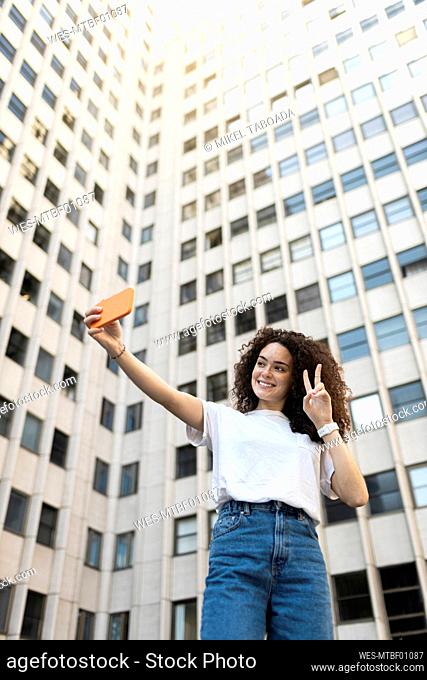 Young woman making peace sign while taking selfie in front of building