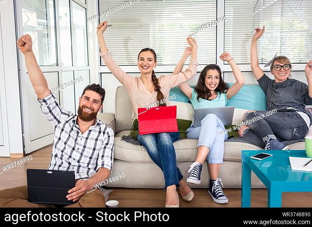 Group portrait of four happy and successful remote workers looking at camera with raised arms while sharing the facilities of a modern co-working space