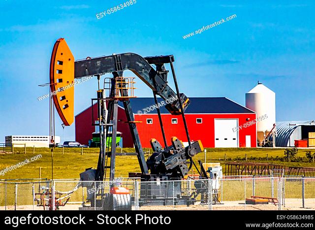 A oil well pumpjack, overground reciprocating pump, is seen by a large ranch in Alberta. With grain silo, red barn and farm buildings in background