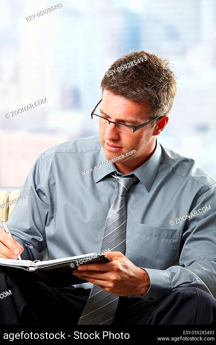 Mid-adult professional with glasses taking notes into organiser sitting on couch