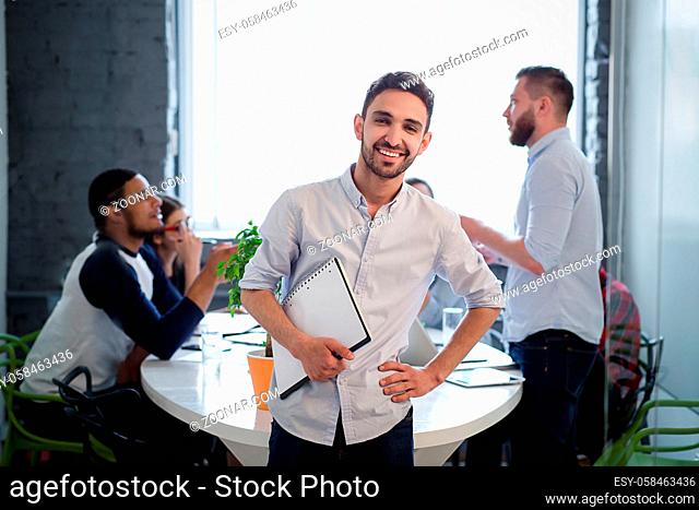 Picture of handsome bearded man in white shirt standing with his hand on hip. Executive businessman posing with clipboard in office interior and smiling