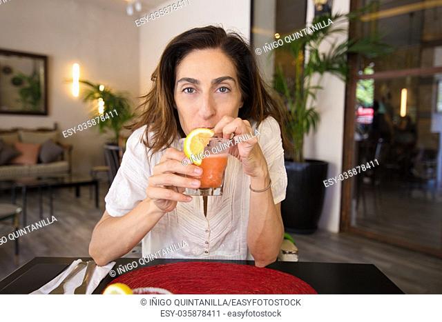 adult woman with white shirt looking and drinking from straws strawberry margarita with slice lemon of glass in hand, sitting in black table with red mat straw...