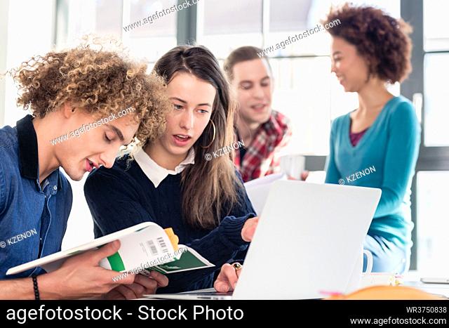 Two young students searching for an answer to their question both online and in a textbook while sitting at desk in the classroom during break
