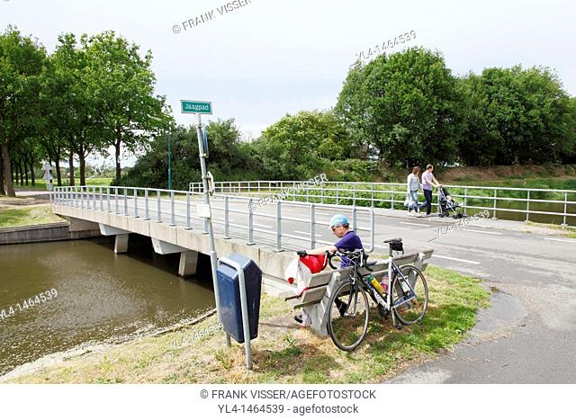 A biker takes a break along a canal while a couple with a buggy walks on the bridge, Netherlands