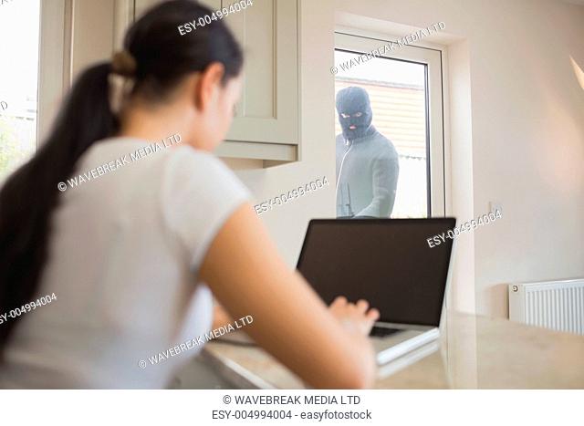 Burglar looking at woman using laptop in kitchen through glass door