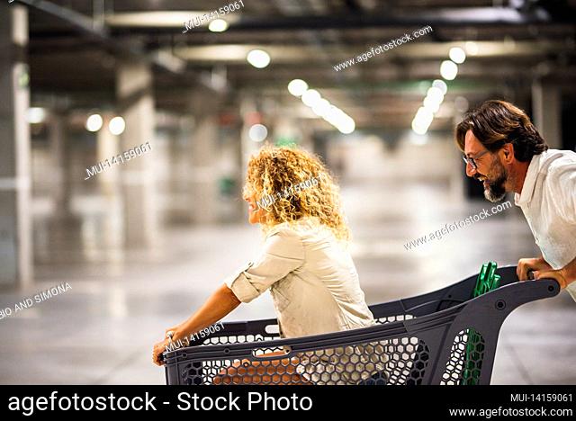 happy couple having fun with shopping cart at parking garage, husband pushing his carefree wife inside shopping trolley. couple is having fun with supermarket...