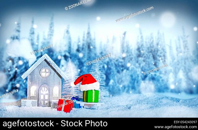 Christmas card with cozy small decorative house , gifts and Santa Claus cap on sleigh on snow over fairy tale winter fir forest background and falling snow