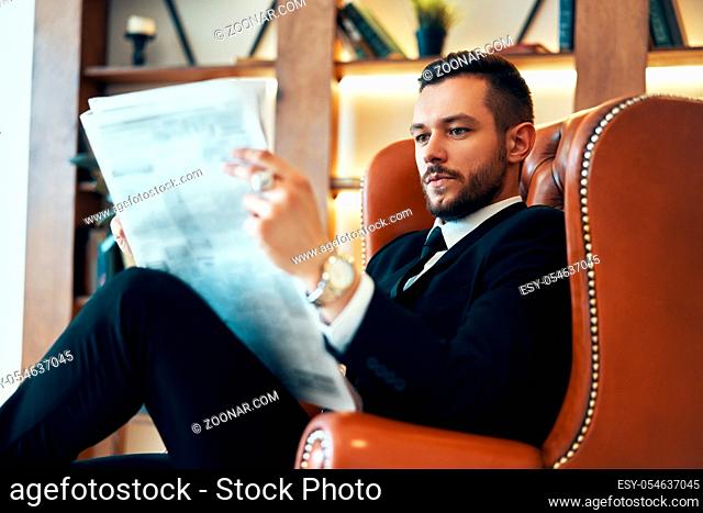 Confident young businessman reading newspaper and latest news while sitting in armchair in the morning in modern interior