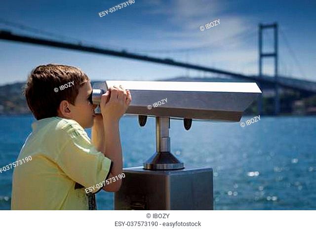Young boy looking through a coin operated viewpoint binoculars at seaside landscape of Istanbul next to a bosphorus bridge