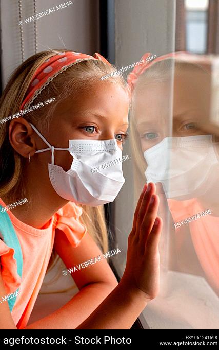 Close-up of caucasian elementary schoolgirl wearing mask while looking through window