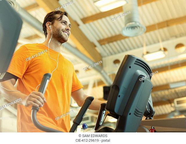 Smiling man with headphones using elliptical trainer at gym