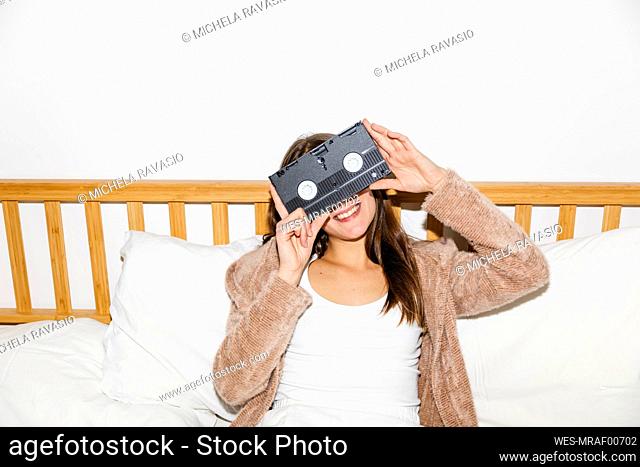 Smiling young woman covering eyes with videocassette in bedroom